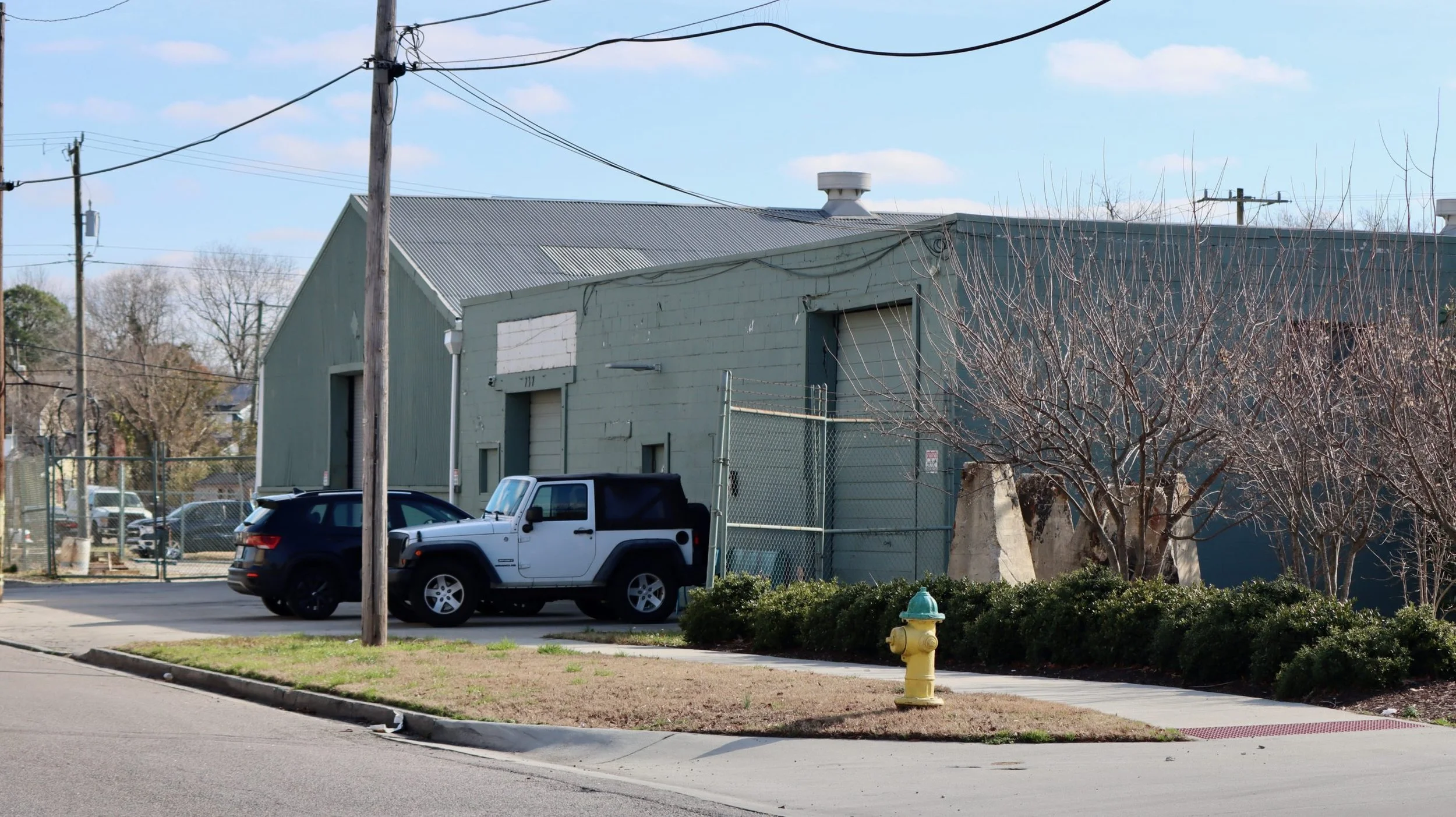 Side view of our Norfolk industrial building with a metal roof, power lines, two parked vehicles, a sidewalk, a yellow fire hydrant, small trees, and a chain-link fence.