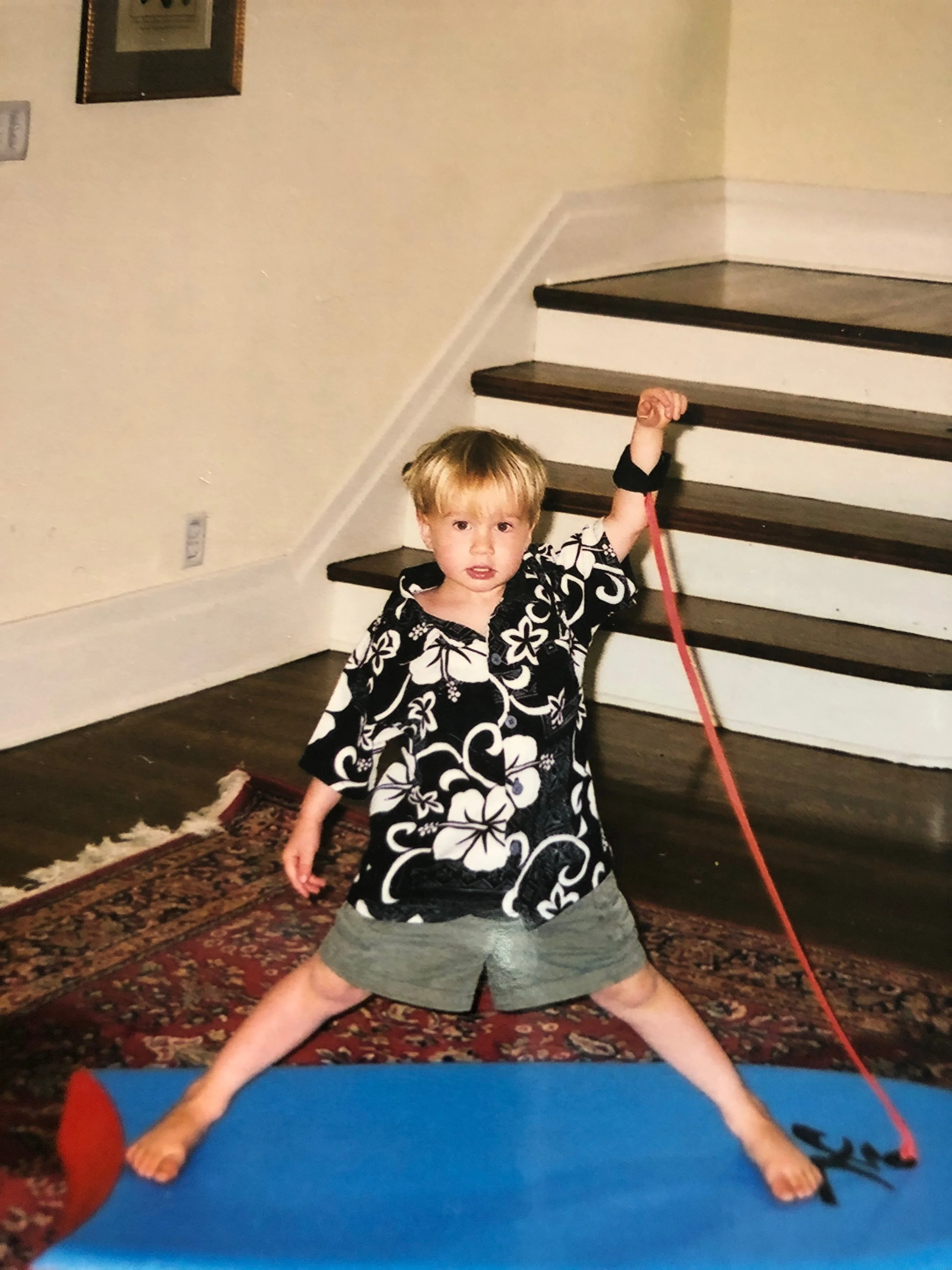 A young boy with blonde hair, wearing a black and white floral shirt and gray shorts, stands indoors on a patterned rug. He is holding a red stick or leash, with legs spread wide, in front of a staircase with dark wooden steps.