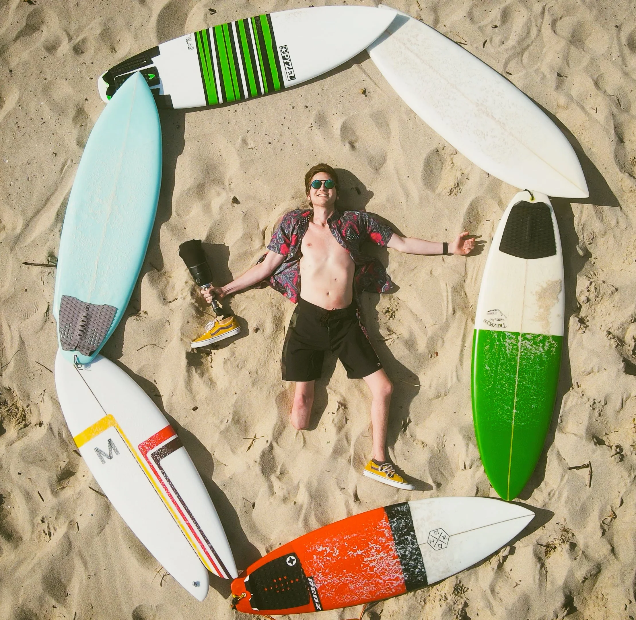 A man lying on sandy beach ground surrounded by seven surfboards in a circle, wearing sunglasses, black shorts, yellow sneakers, and an open shirt, holding a prosthetic leg, smiling, with sunlight casting shadows.
