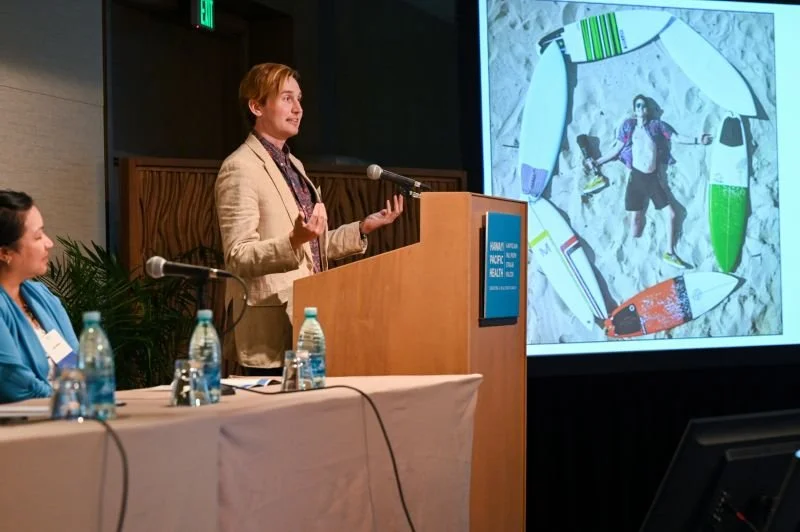 Woman presenter at a conference giving a speech at a podium with a large screen behind displaying a photo of a snowboard with various items on snow.