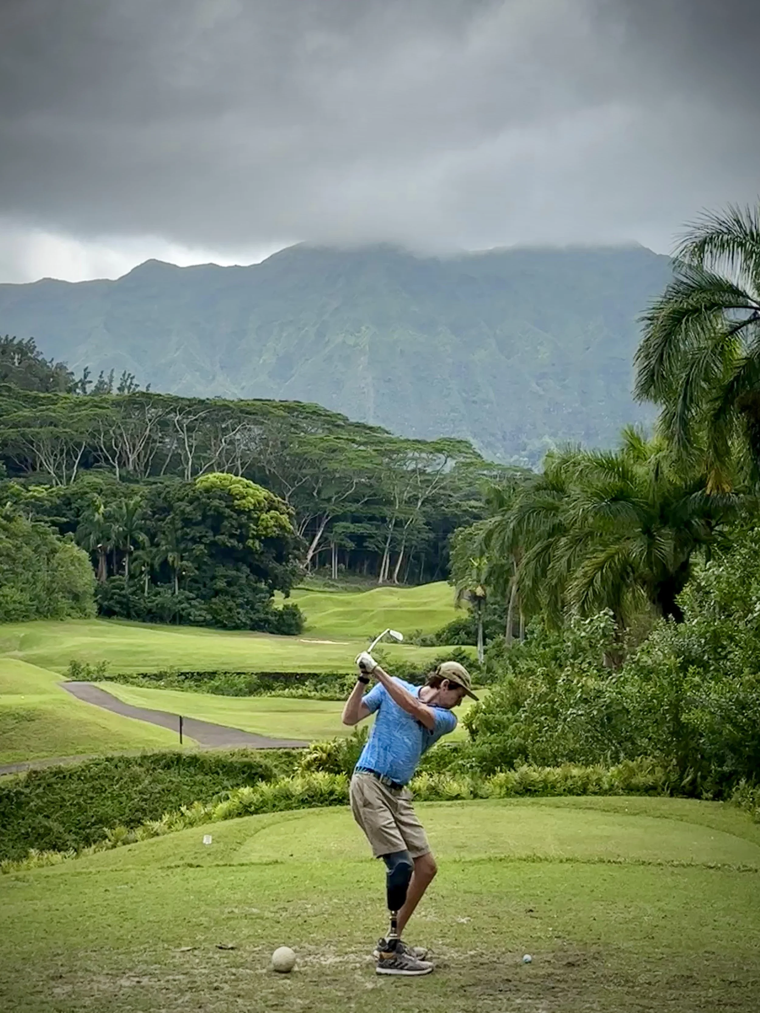 A man in a blue shirt and beige shorts teeing off on a golf course surrounded by lush green trees and mountains in the background, under a cloudy sky.