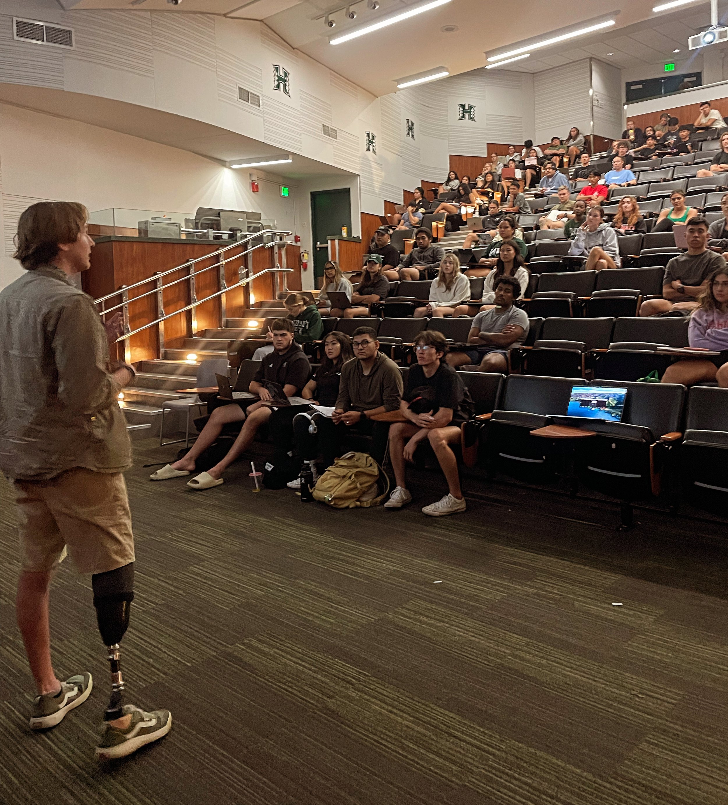 A person with a prosthetic limb giving a presentation or lecture to a seated audience in a tiered auditorium or lecture hall.