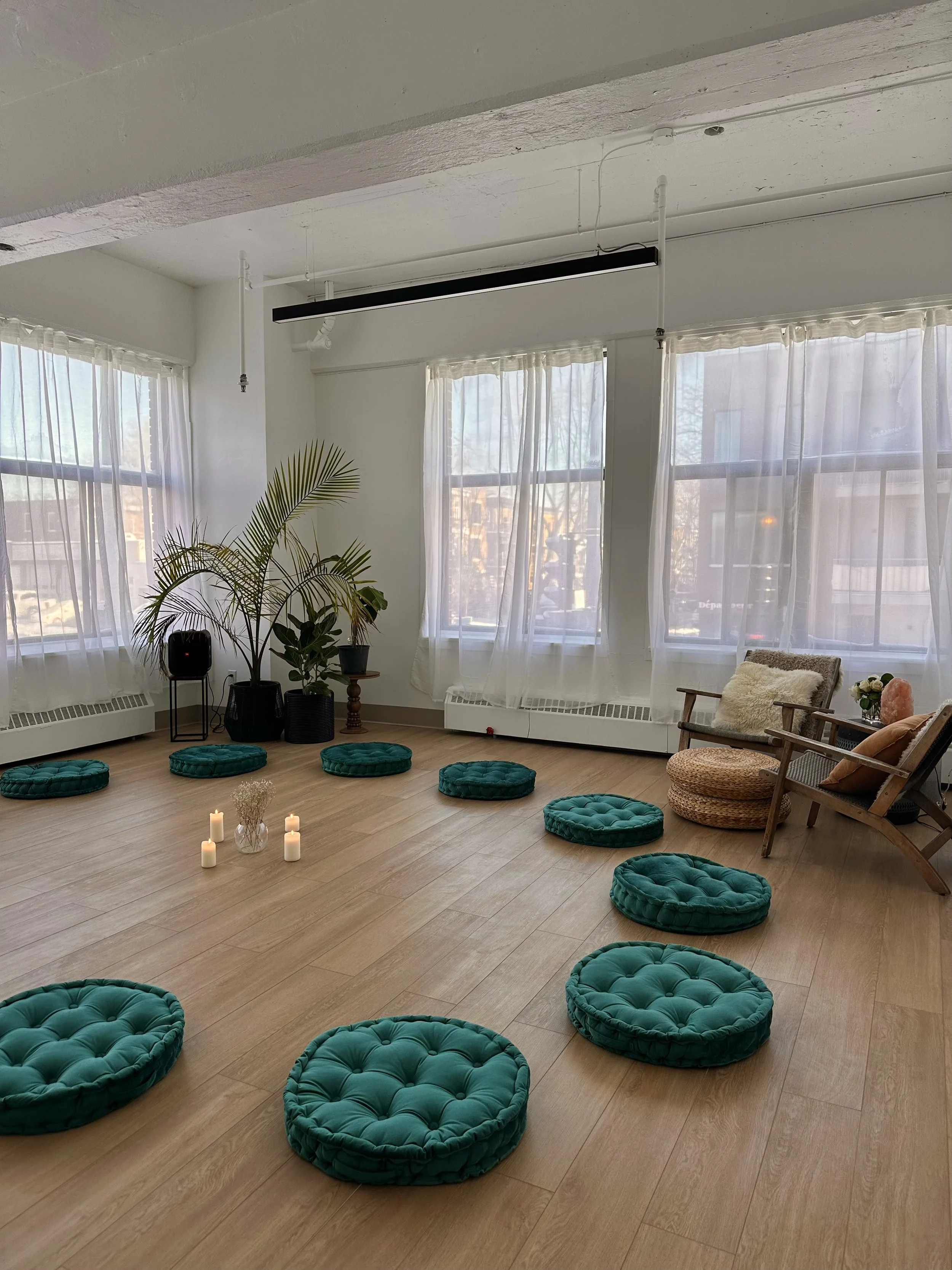 A cozy, well-lit meditation or yoga room with eight round teal cushions arranged on a wooden floor in a circle, a small table with candles and dried flowers in the center, large windows with white curtains, a few potted plants, and a comfortable chair with cushions and a side table on the right.