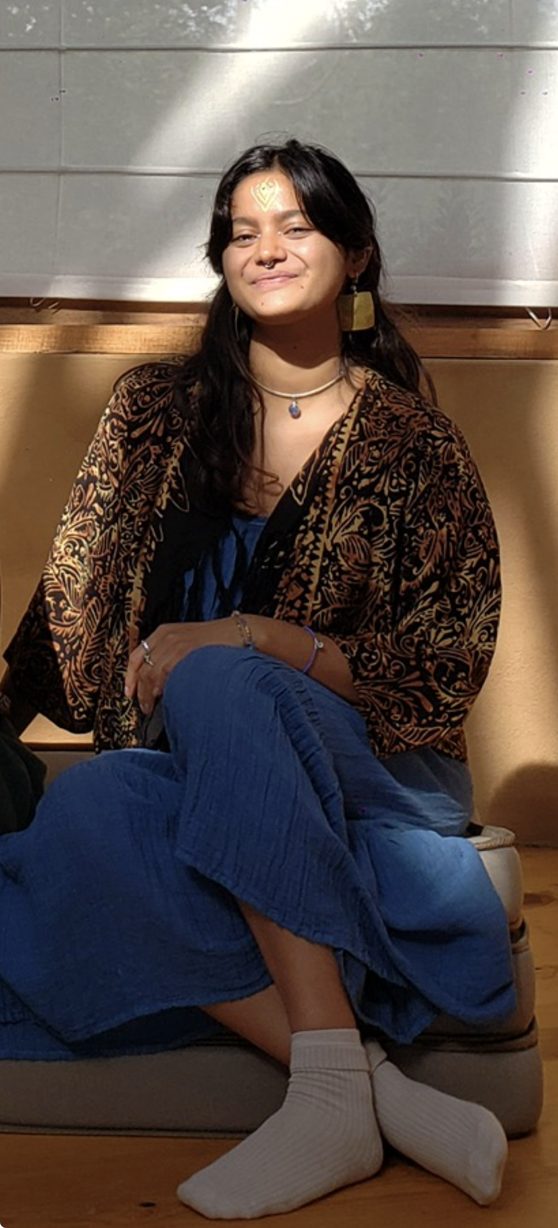 Young woman with dark hair and a septum piercing, sitting indoors, smiling, wearing a patterned jacket, blue jeans, and white socks.