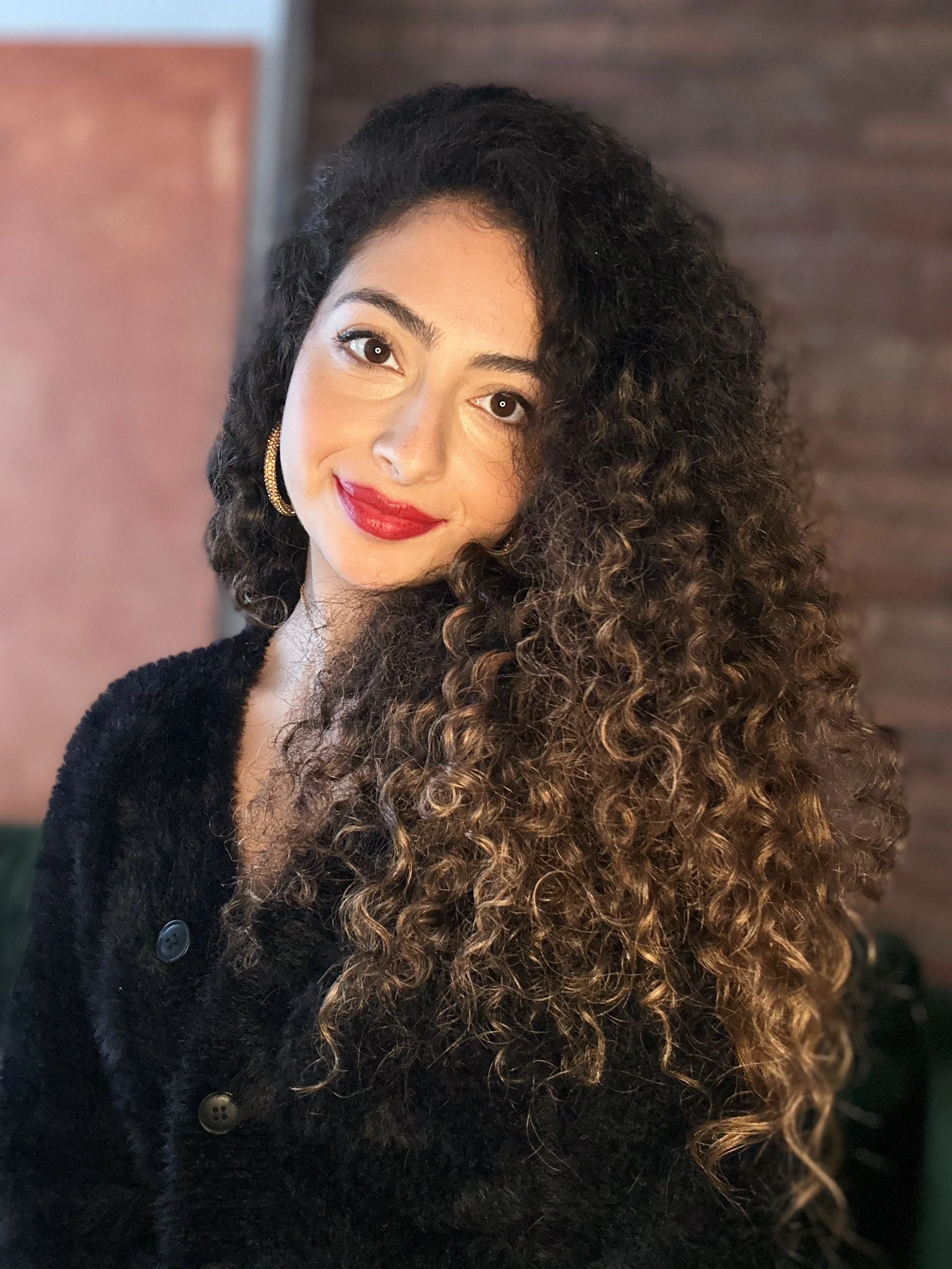 A woman with curly brown hair wearing a black coat with large buttons and gold hoop earrings.