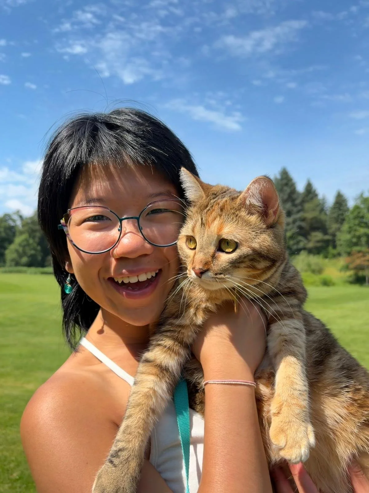 A young woman smiling and holding a tabby cat outside on a sunny day with green trees and blue sky in the background.