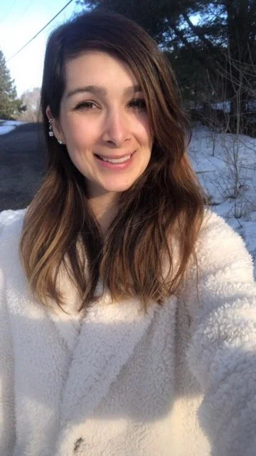 Young woman taking a selfie outdoors, smiling, with snow on the ground and trees in the background.