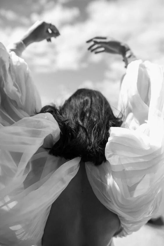 Back view of a woman with dark hair wearing flowing white fabric, possibly a dress or robe, with her arms raised.