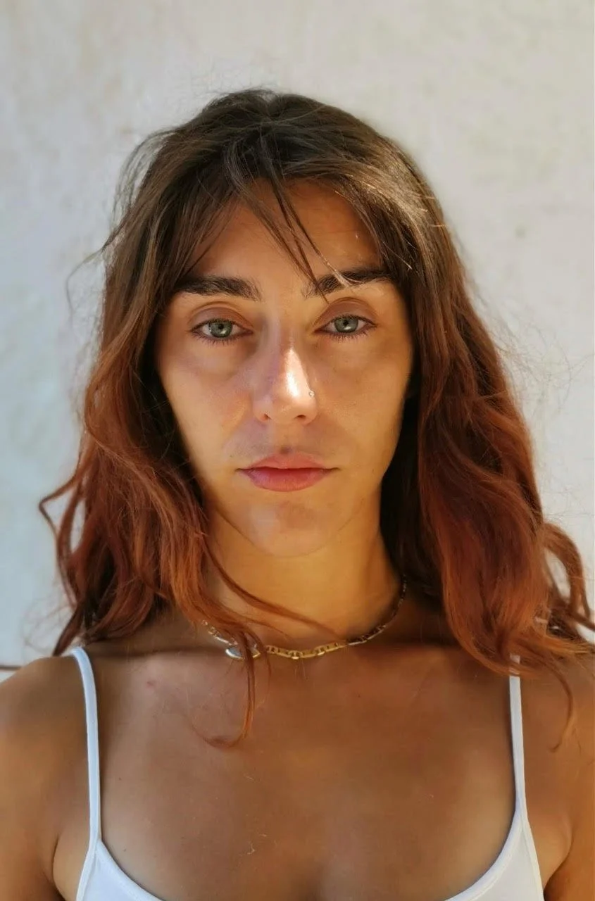 Close-up of a woman with light skin, green eyes, shoulder-length wavy brown hair, wearing a white spaghetti strap top and a gold chain necklace, standing against a plain light background