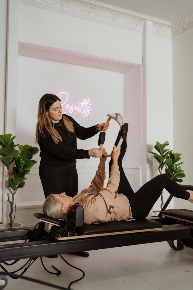 A Pilates instructor in black assisting an older woman in a pink hoodie in a Pilates or physical therapy session on a reformer machine, with a neon sign reading 'Results' in the background and green potted plants on either side.