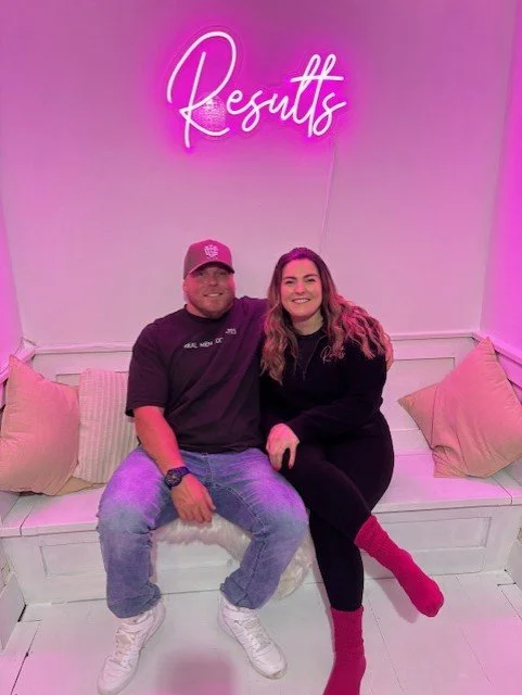 A man and woman sitting on a white bench with pillows, smiling, in a room with pink lighting and a neon sign that says 'Results' above them.