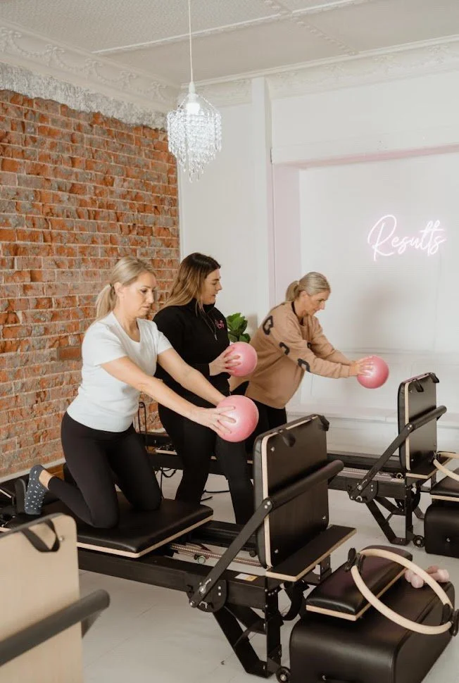 Three women participating in a Pilates class using reformer machines, each holding pink exercise balls, in a room with a brick wall and a chandelier.