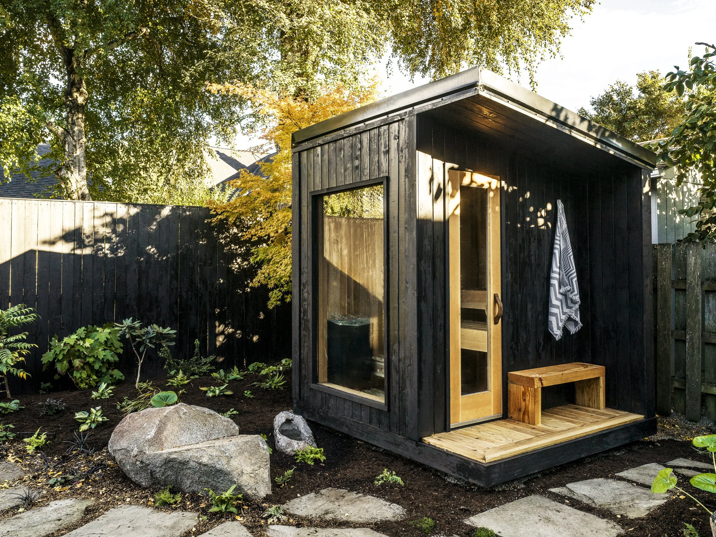 A small backyard sauna with black wooden exterior, a glass door, and large window, surrounded by a garden with plants, large rocks, and stepping stones, fencing, and trees in the background.