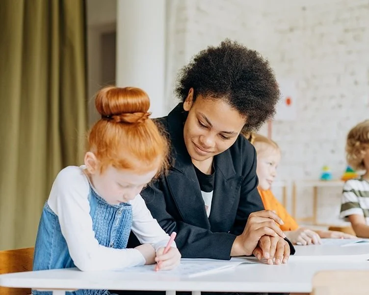 A teacher with natural curly hair helping a young girl with red hair and a top knot with her schoolwork in a classroom with other children in the background.