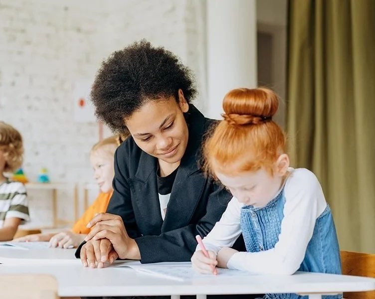 A woman with curly hair and a black blazer sitting next to a red-haired girl in a denim dress writing on a piece of paper, with other children in the background in a classroom.