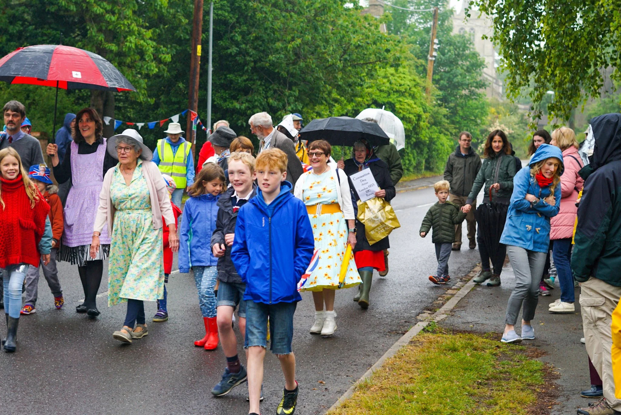 A group of people, including children and adults, walking outdoors in rainy weather, some holding umbrellas, on a street surrounded by green trees.