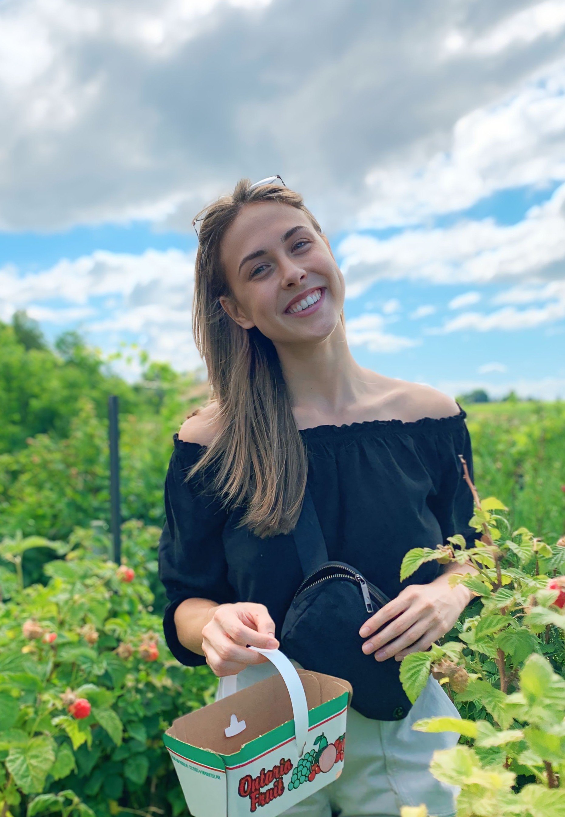 A smiling woman picking strawberries in a green field on a partly cloudy day.