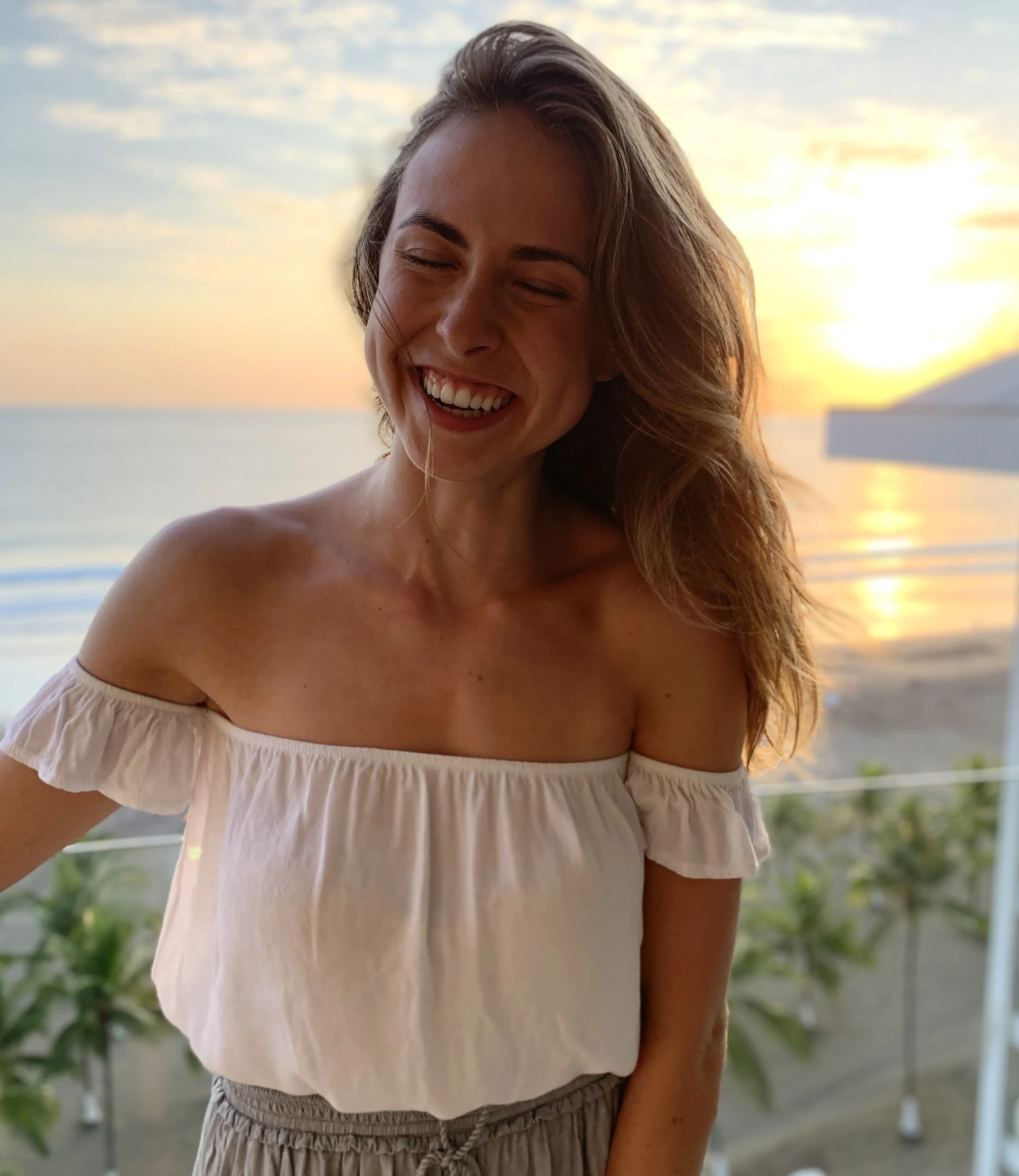 A woman with long wavy hair smiling and laughing at sunset on a beach, wearing an off-the-shoulder white top and light-colored pants.