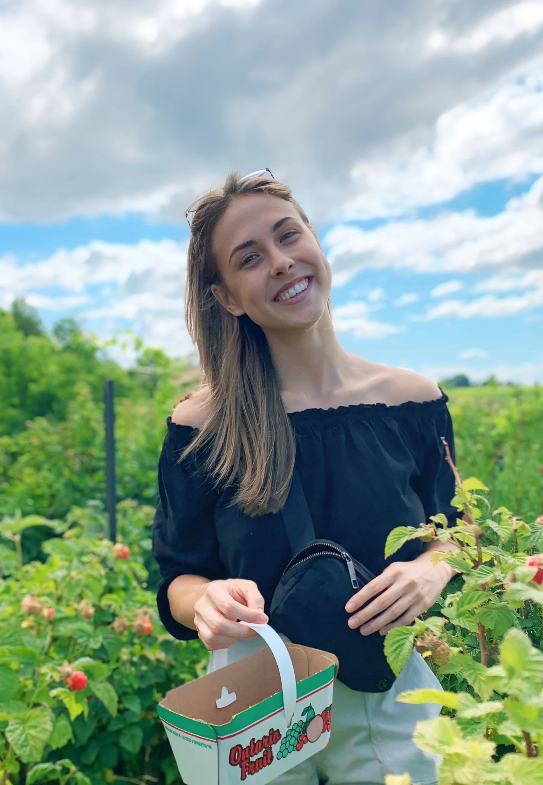 A smiling young woman in a black off-shoulder top holding a basket of raspberries outdoors in a raspberry field with green plants and a partly cloudy sky in the background.