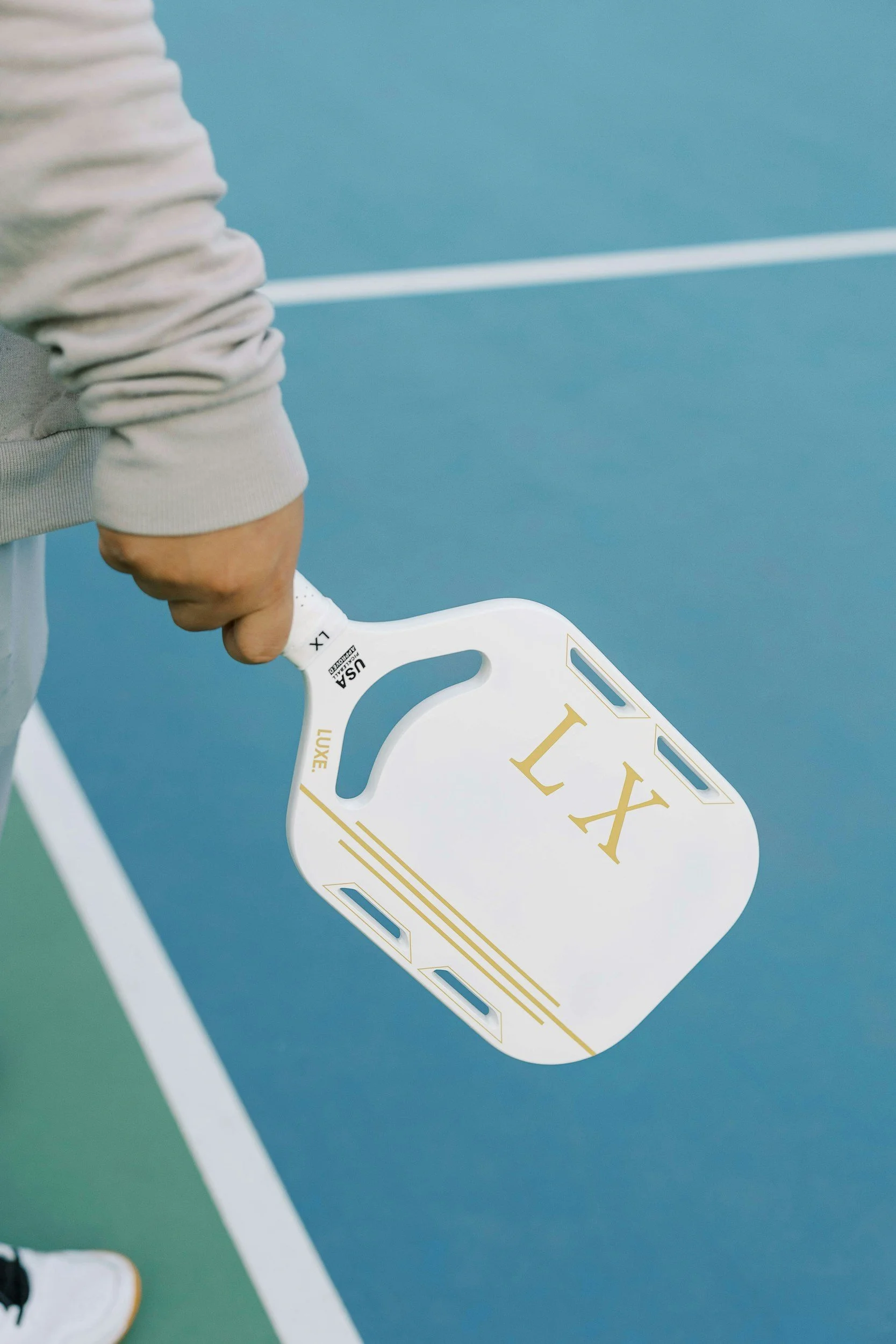 Close-up of a person holding a tennis racket with a white frame and gold accents on a tennis court with blue and green surface.