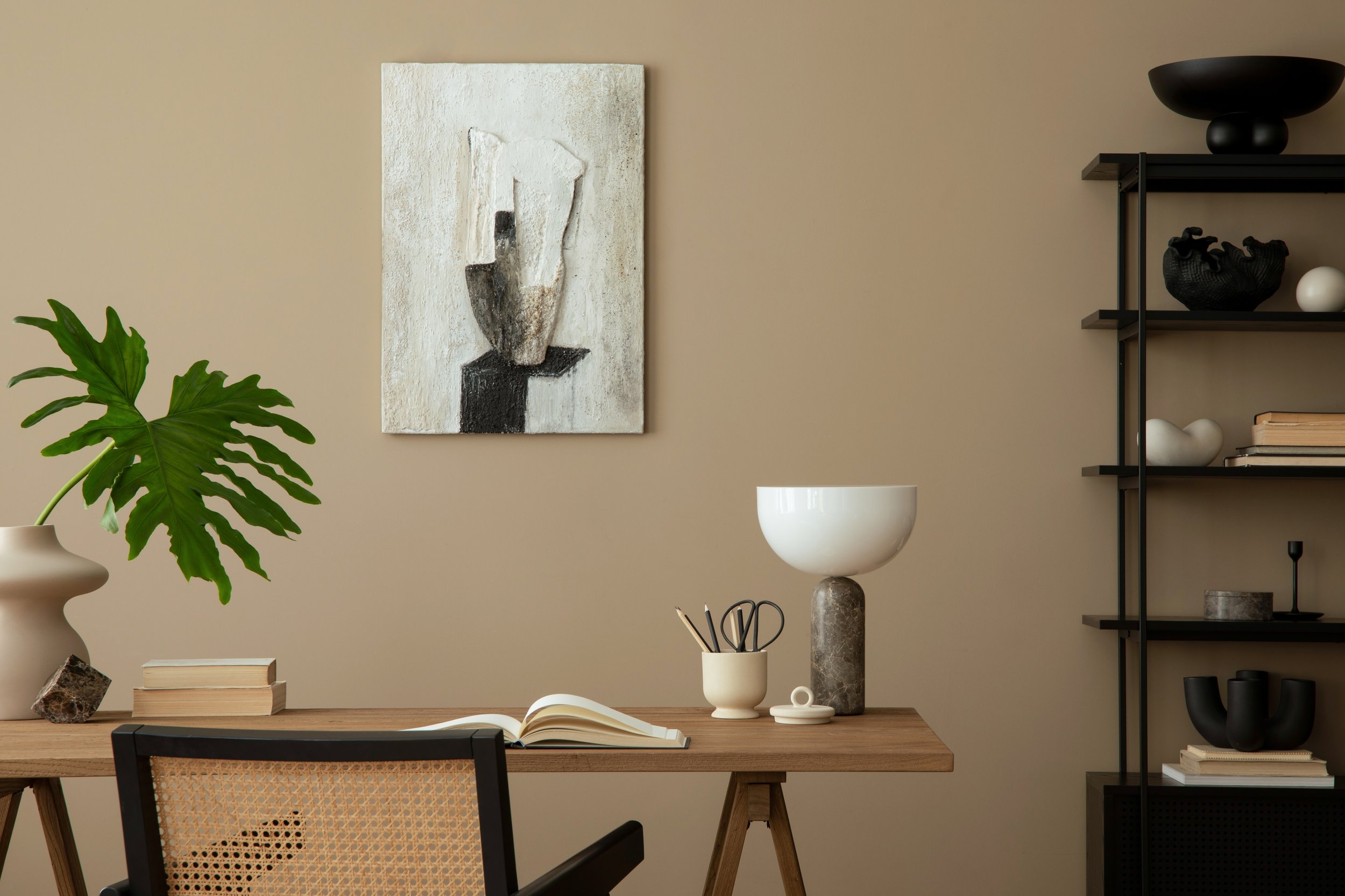 A modern workspace with a wooden desk, a black and beige chair, a large green leafy plant, a beige wall art piece, and a black shelving unit with decorative objects.
