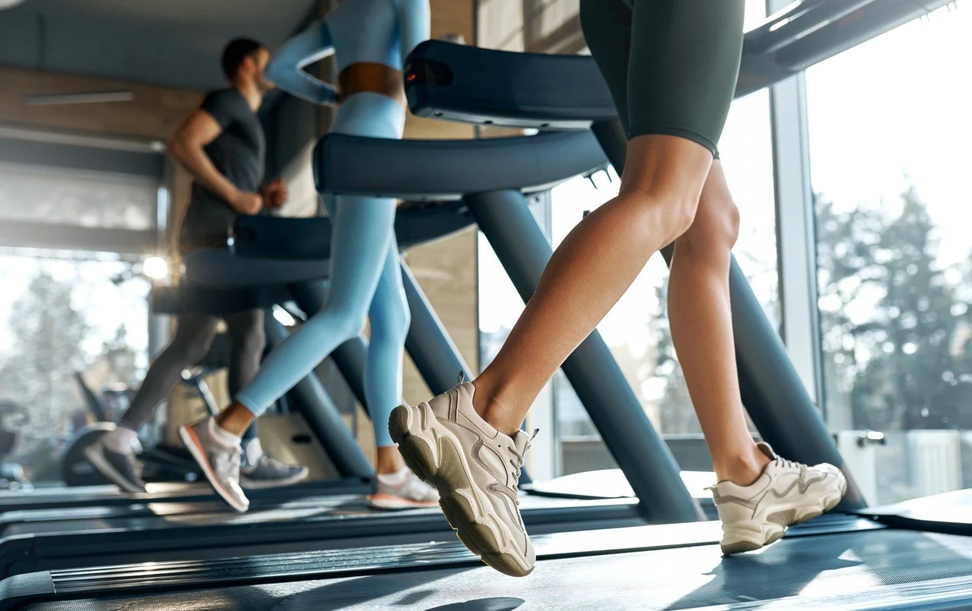 People running on treadmills in a gym with large windows and sunlight.