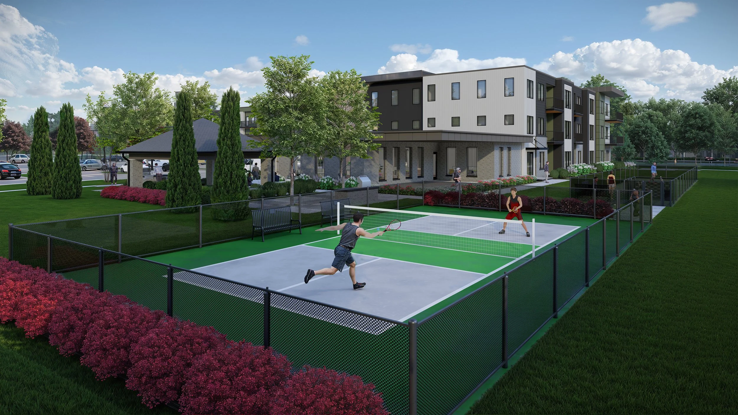 People playing pickleball on an outdoor court surrounded by a black fence in a residential community with a modern apartment building, trees, shrubs, and a parking lot in the background.