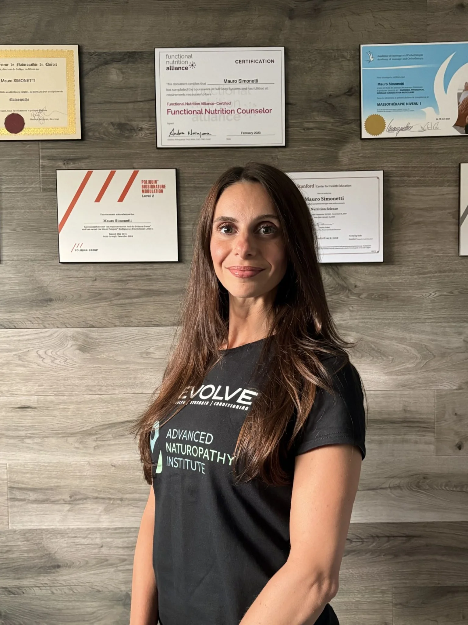 A woman standing indoors with a wall of framed diplomas and certificates behind her, wearing a black T-shirt that says 'EVOLVE' and 'ADVANCED NATUROPATHY INSTITUTE'.