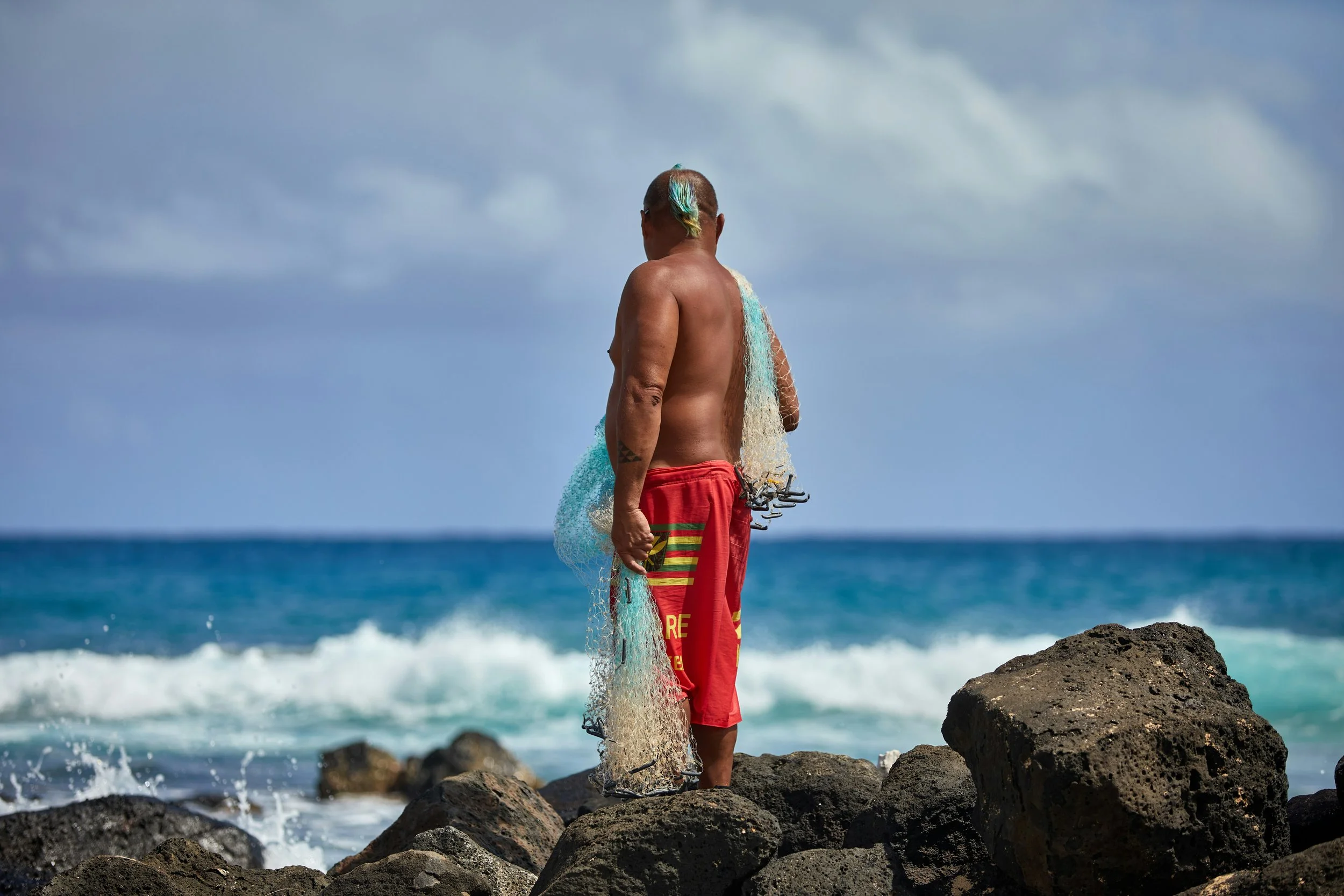 A man with a dyed mohawk hairstyle, shirtless, wearing red shorts, standing on rocks near the ocean, holding fishing gear and a fishing net, facing away from the camera with waves in the background.