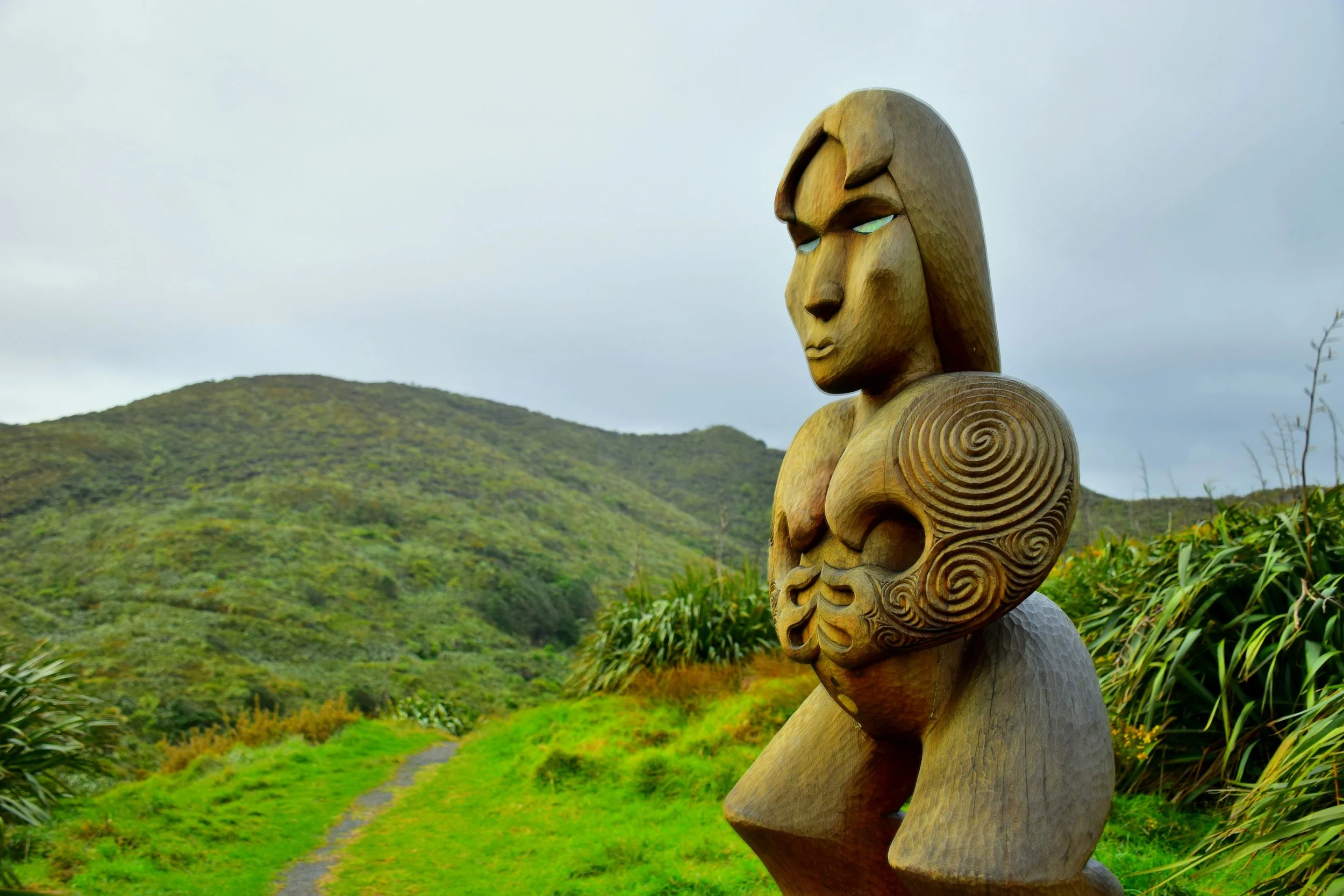 Wooden Maori-style sculpture of a female figure with stylized hair and body motifs in a lush green landscape with a mountain and cloudy sky in the background.