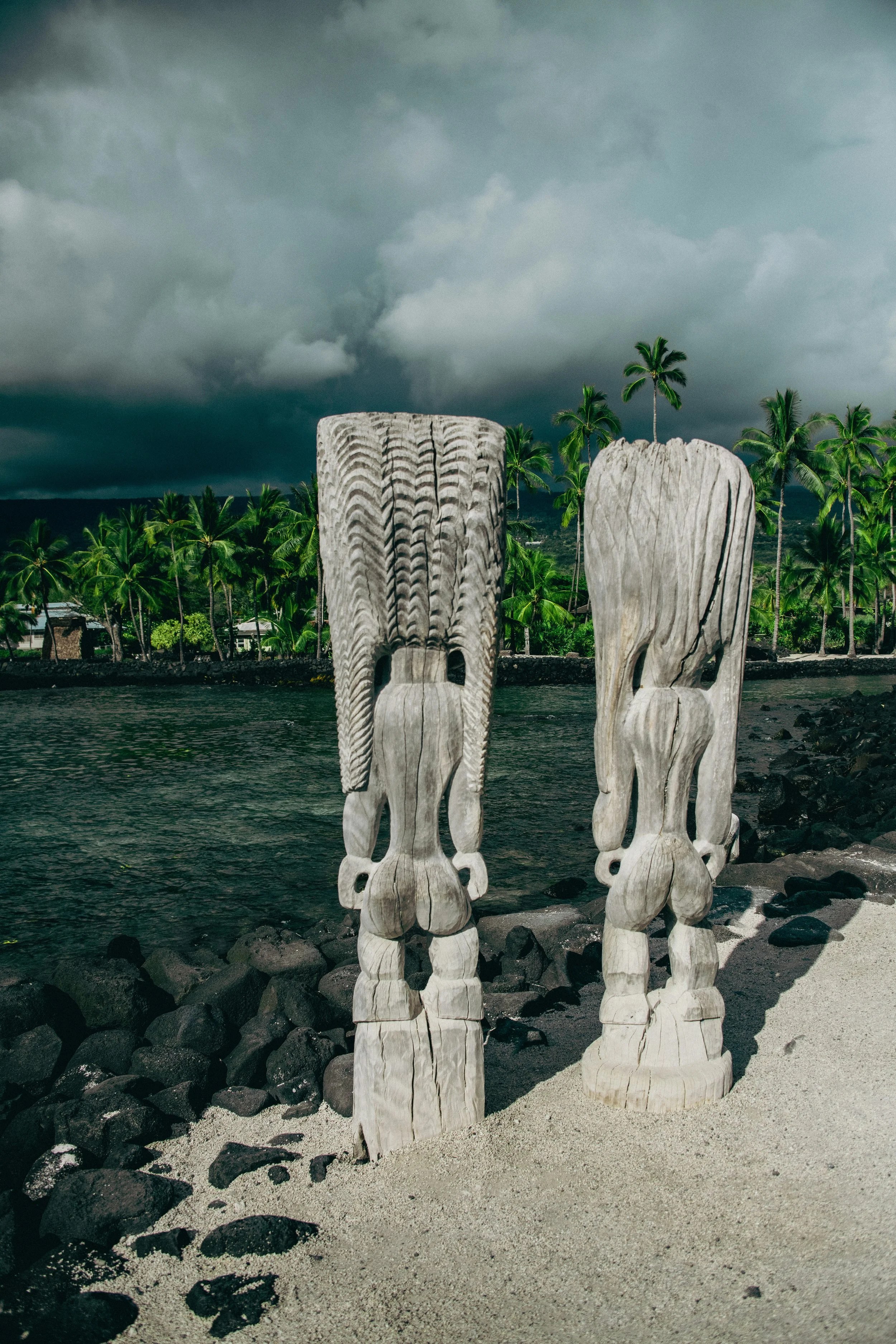 Two carved wooden Tiki statues on a sandy beach with a rocky shoreline, palm trees, and dark storm clouds in the background.