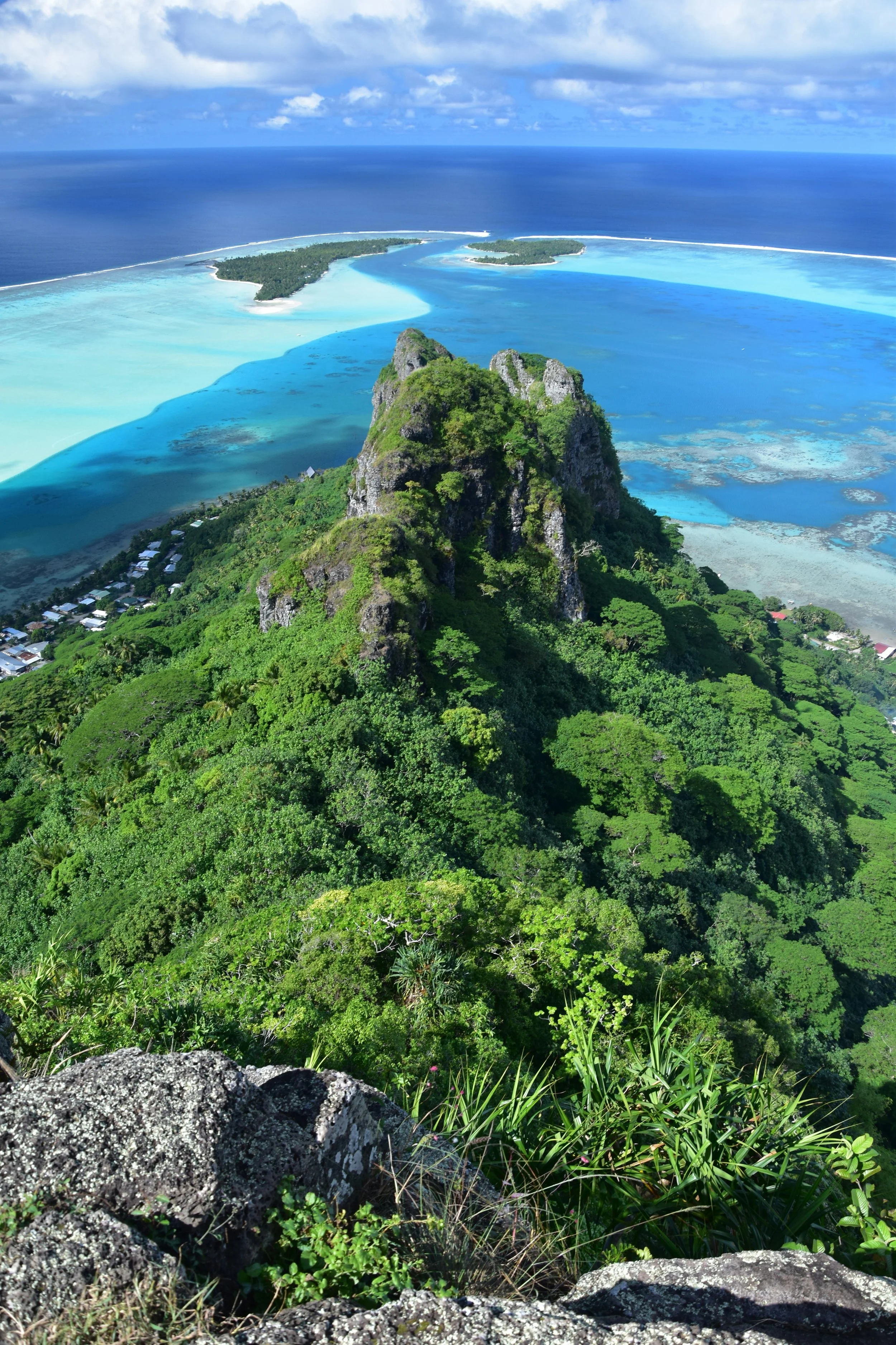 Aerial view of a lush green mountain with rocky peaks overlooking clear blue ocean waters and an island with sandy beaches in the distance under a partly cloudy sky.