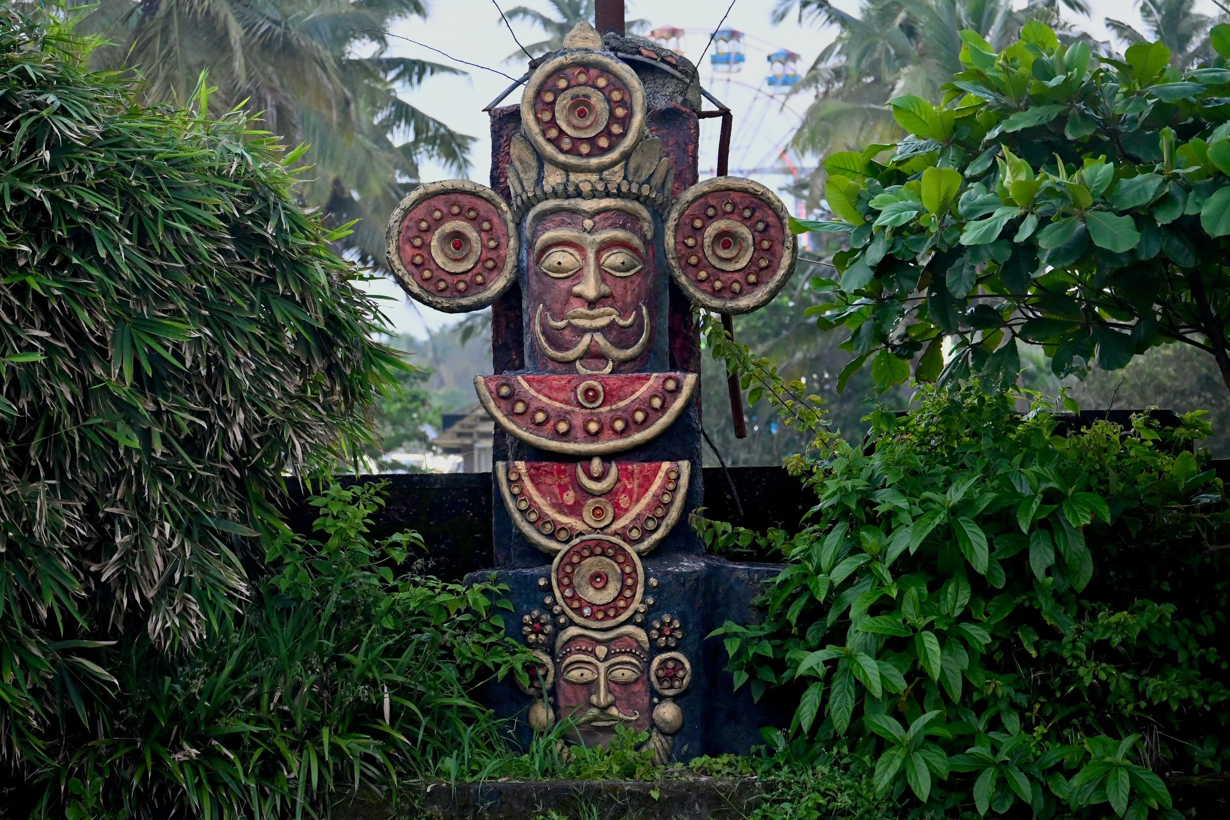 Colorful tribal statue of a face with elaborate circular decorations, surrounded by green bushes and trees in an outdoor setting.