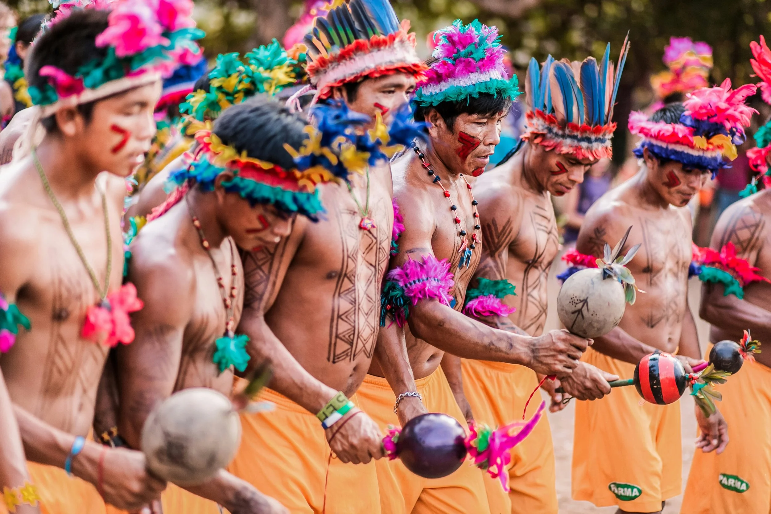 Group of indigenous men dressed in traditional attire with feathered headdresses, painted faces, and holding ceremonial staffs, participating in a cultural dance or ceremony.