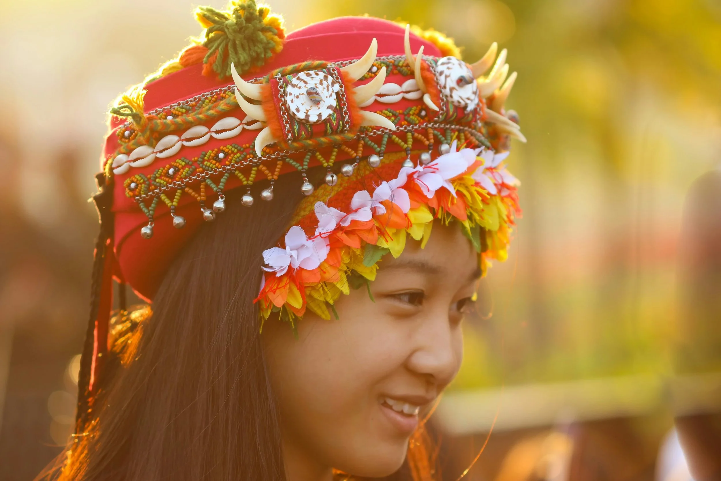 Young woman wearing traditional tribal headdress decorated with shells, beads, chains, and colorful flowers, smiling outdoors during sunset.