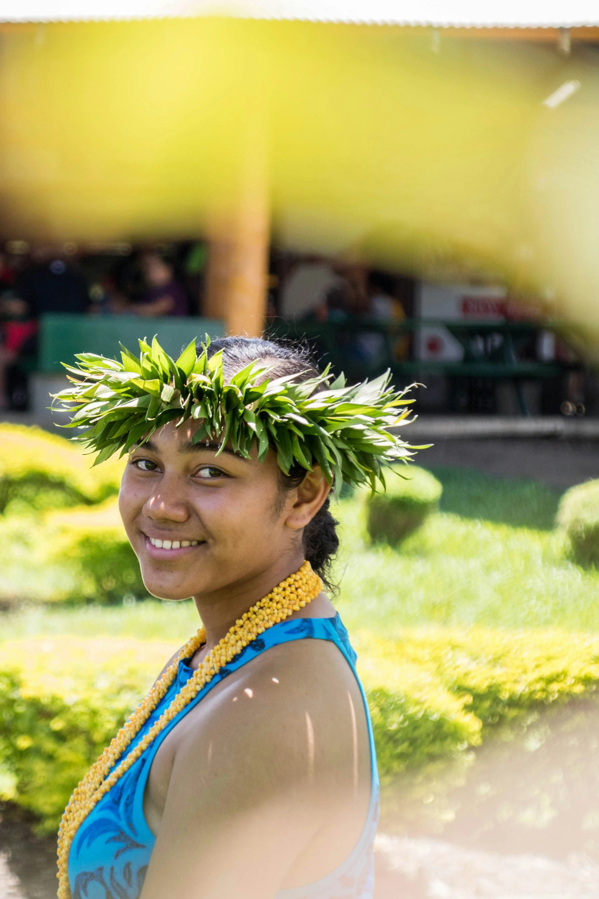 Young woman smiling at a cultural event, wearing a blue dress, a necklace of yellow beads, and a leafy headdress.