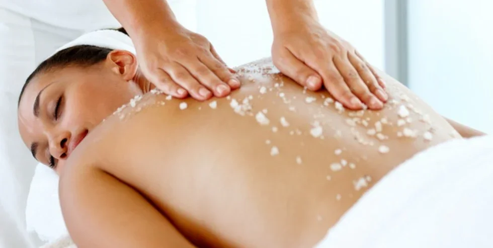 A woman receives a back massage with salt crystals sprinkled on her back, lying face down in a massage setting.