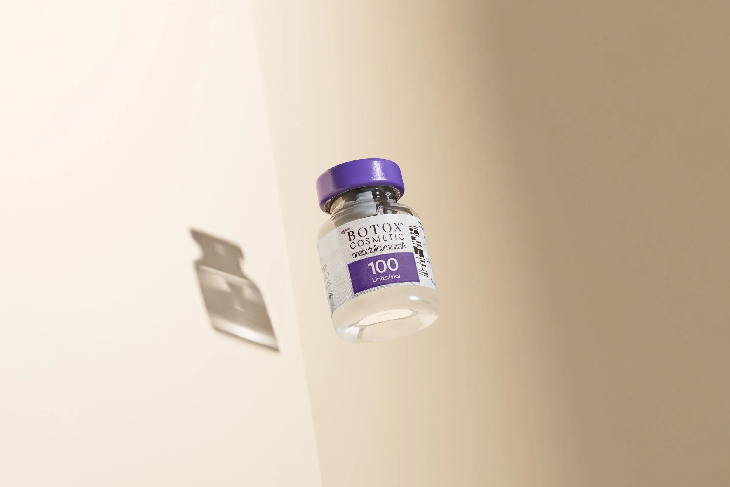 A small vial labeled 'Botox Cosmetic' with a purple cap on a plain surface, casting a shadow.