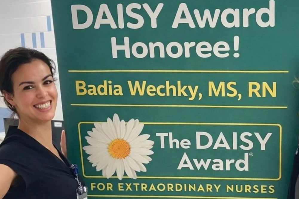 A woman standing next to a sign that reads "DAISY Award Honoree! Badia Wechky, MS, RN, The DAISY Award for extraordinary nurses," with a picture of a daisy flower on it.