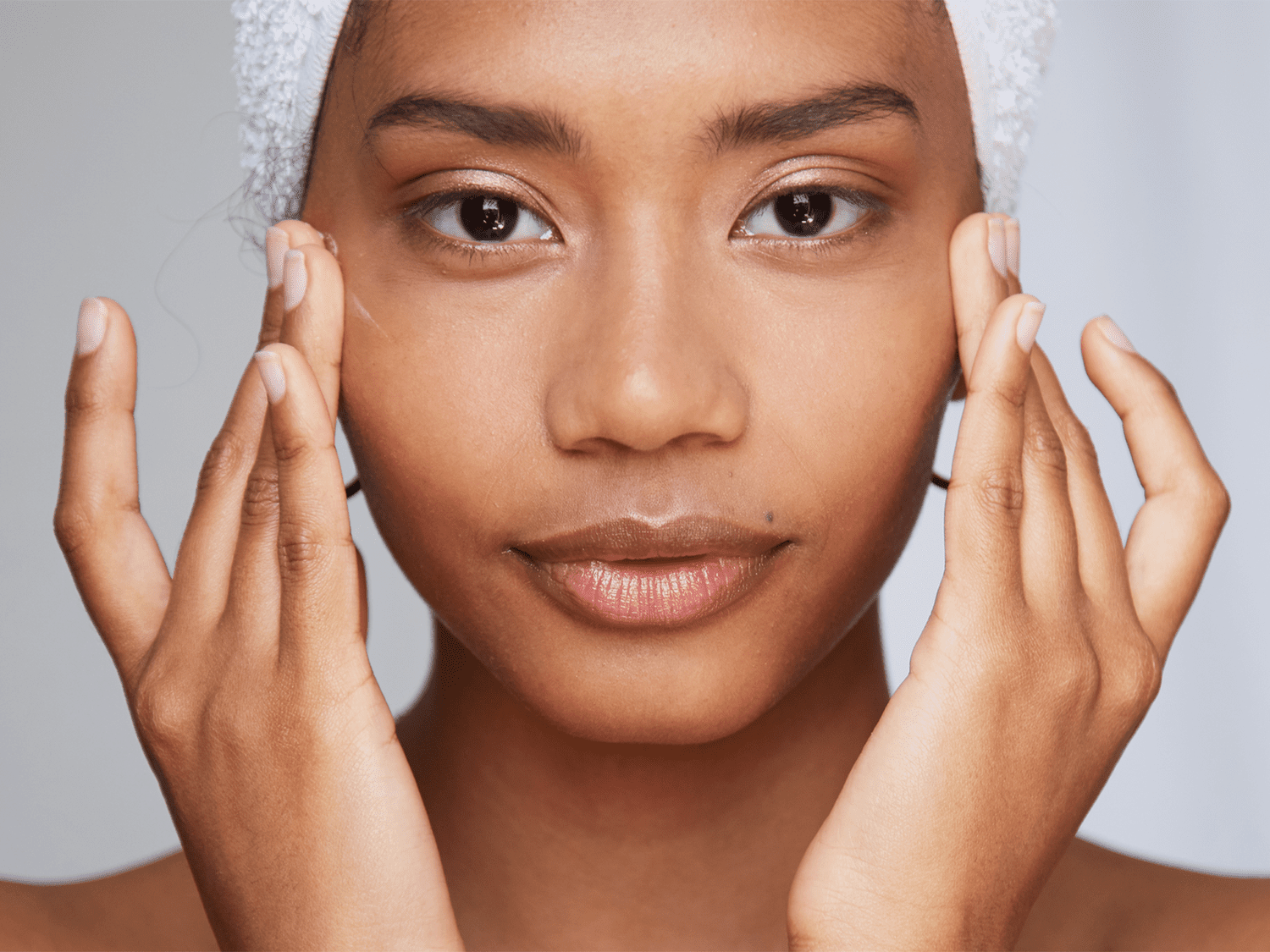 Close-up of a woman with smooth skin, gently touching her cheeks with both hands, against a neutral background.
