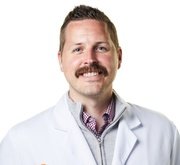 Portrait of a man with a mustache wearing a white coat and a collared shirt, smiling at the camera.