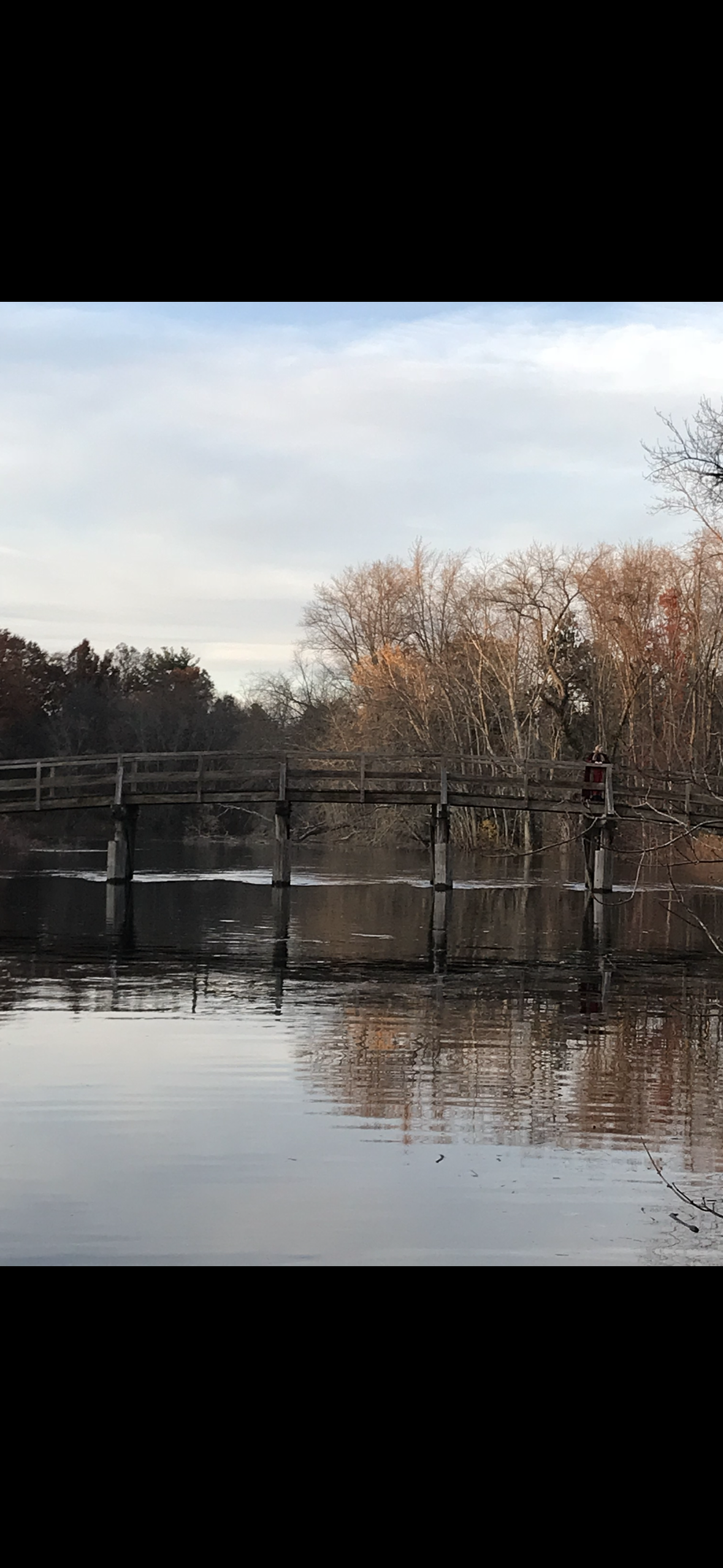 A wooden bridge over a calm river with bare trees and some leaves in fall colors, reflecting in the water, under a partly cloudy sky.