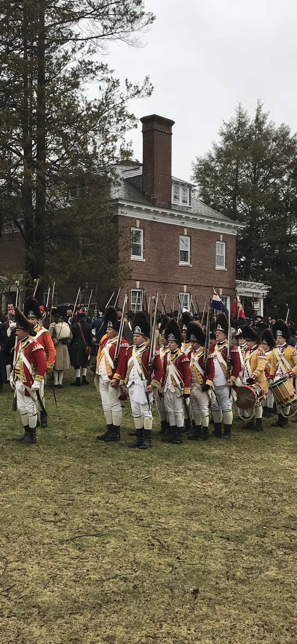 A reenactment of soldiers in 18th-century military uniforms standing on a lawn in front of a brick house, with other participants and spectators in the background, surrounded by trees.