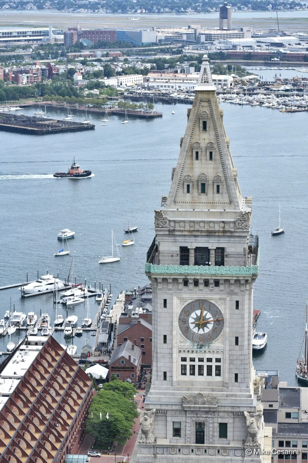 A tall clock tower overlooking a harbor with sailboats and yachts, and a cityscape in the background.