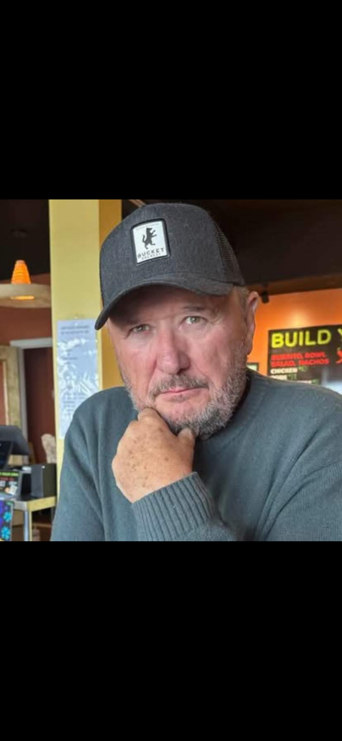 A middle-aged man with a beard and mustache wearing a gray cap with a bear logo and a gray sweater. He is sitting in a restaurant or cafe, resting his chin on his hand, looking at the camera.