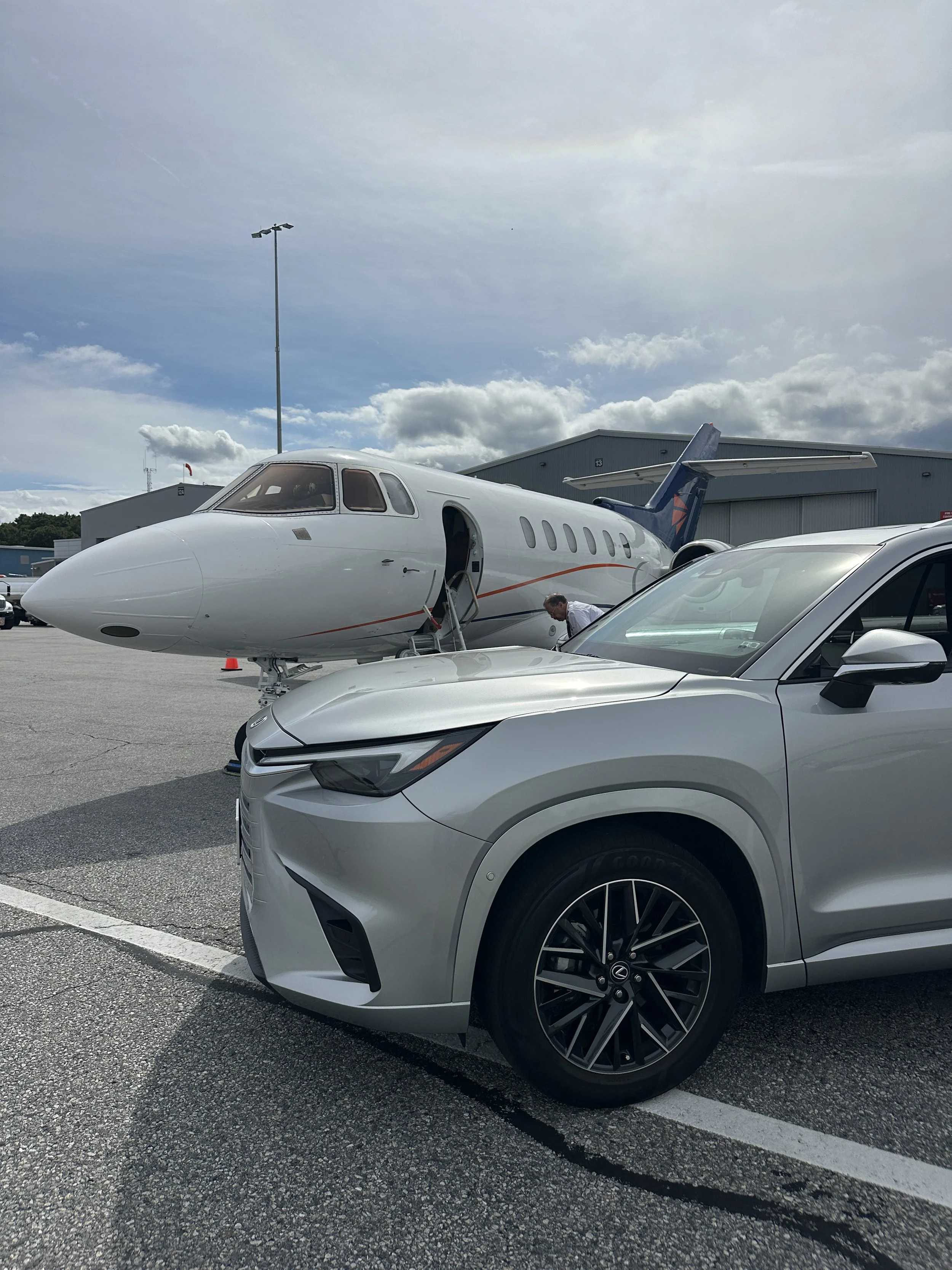 A private jet parked on the tarmac with a man in a white shirt near the staircase and a silver sports car in the foreground. A large hangar is in the background under a partly cloudy sky.
