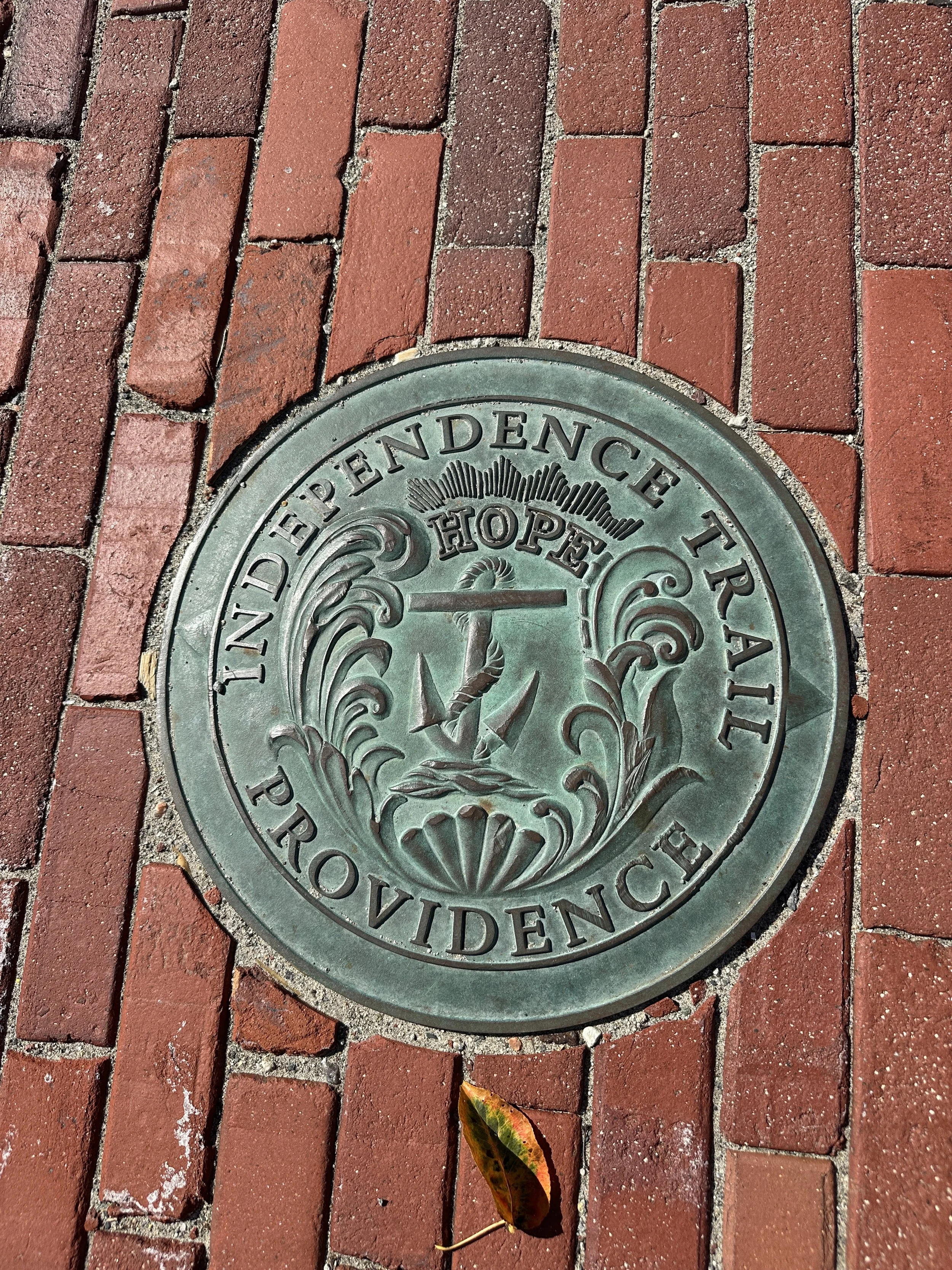 A circular metal plaque embedded in red brick pavement. The plaque reads 'Town of Pendence Trail Providence' with decorative elements. There is a fallen leaf on the pavement near the plaque.