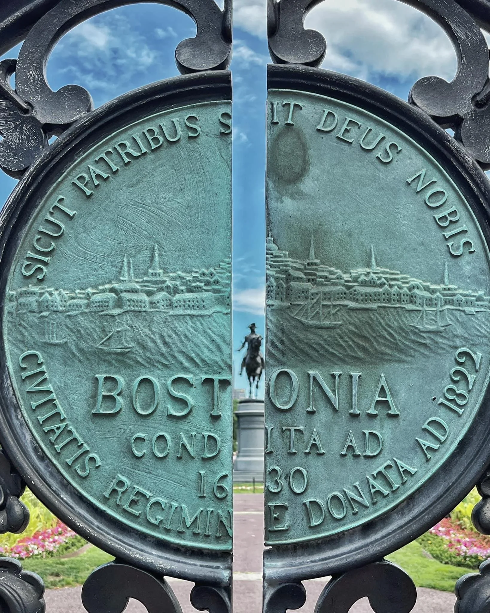 Close-up of two joined metal commemorative coins with engraved cityscape, viewed through decorative black metal fence with a cloudy sky in the background.