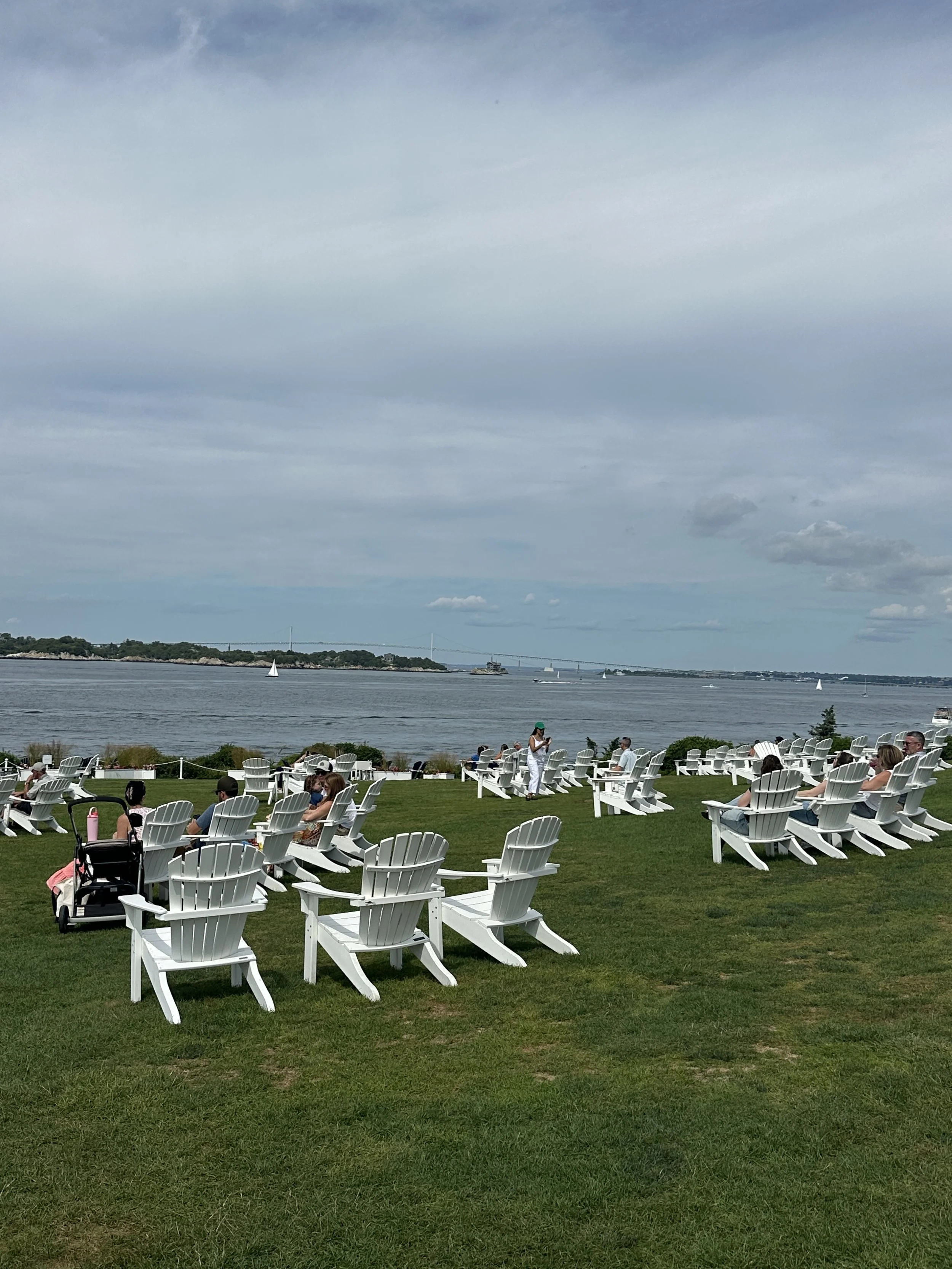 People sitting and standing on a grassy outdoor area near a body of water with sailboats, trees, and a bridge in the background under a partly cloudy sky.