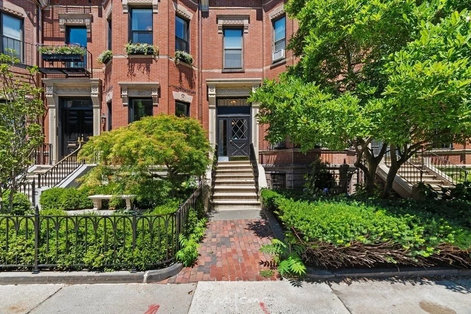 Front view of a red brick townhouse with black doors and stairs, surrounded by green trees and shrubs, black metal fencing, and a small brick pathway leading to the entrance.